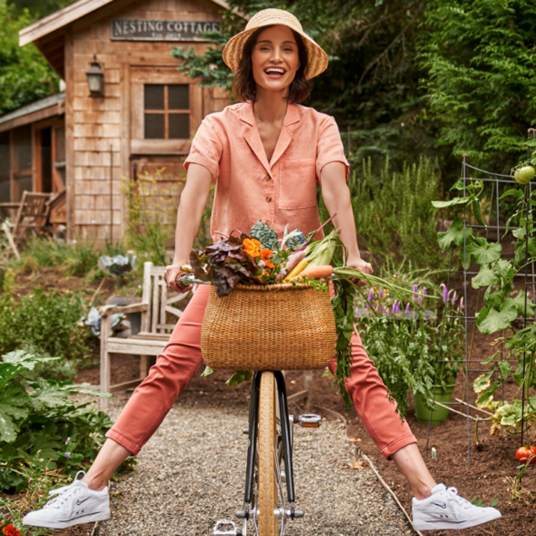 Une femme sur un vélo dans un potager. Le panier du vélo est rempli de légumes frais.