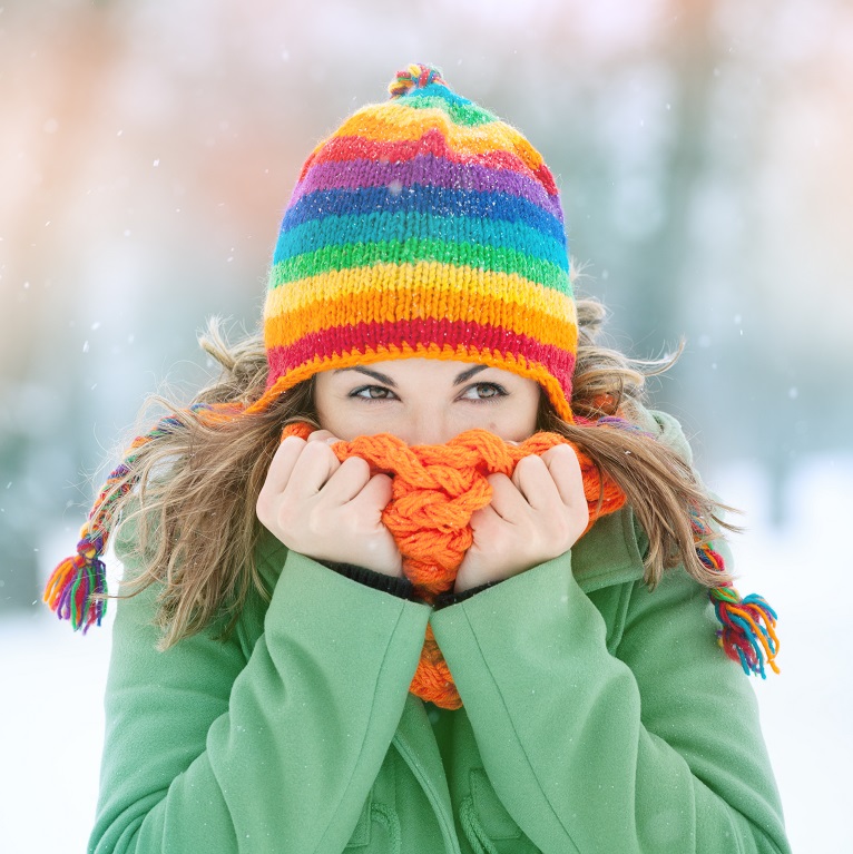 A woman in a green coat and rainbow stocking cap holds her orange scarf over her mouth and nose while trying to stay warm in winter weather.