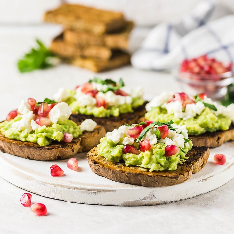 A plate of toast topped with avocado mixed with Nutrilite Organics Green Superfood Powder, pomegranate seeds and feta.