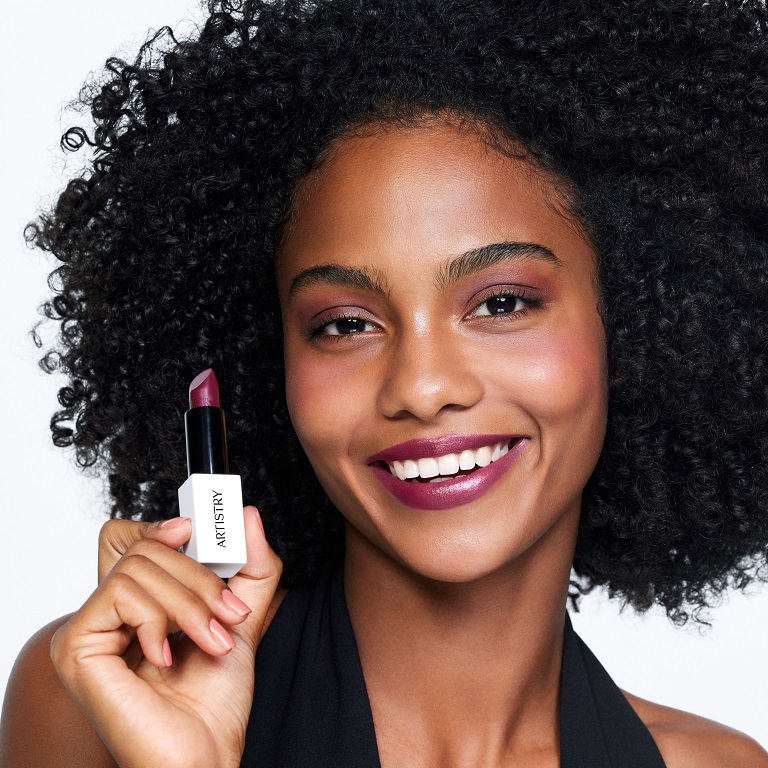 A smiling woman with flawless lips looks at the camera while holding a tube of Artistry Go Vibrant Lipstick.