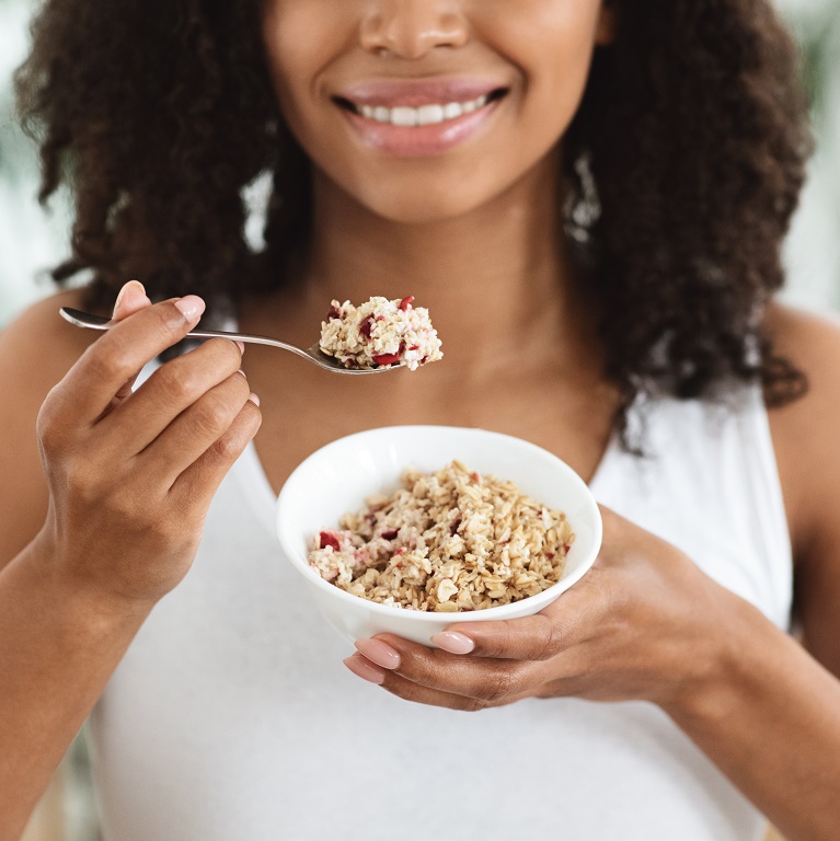 A smiling woman holds a bowl of oatmeal in one hand while raising a spoonful of it up to her mouth.