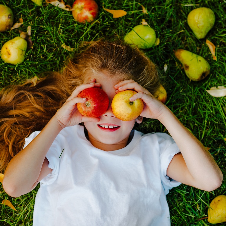 An overhead view of a young girl lying in the grass surrounded by apples and pears. She is holding an apple over each eye.