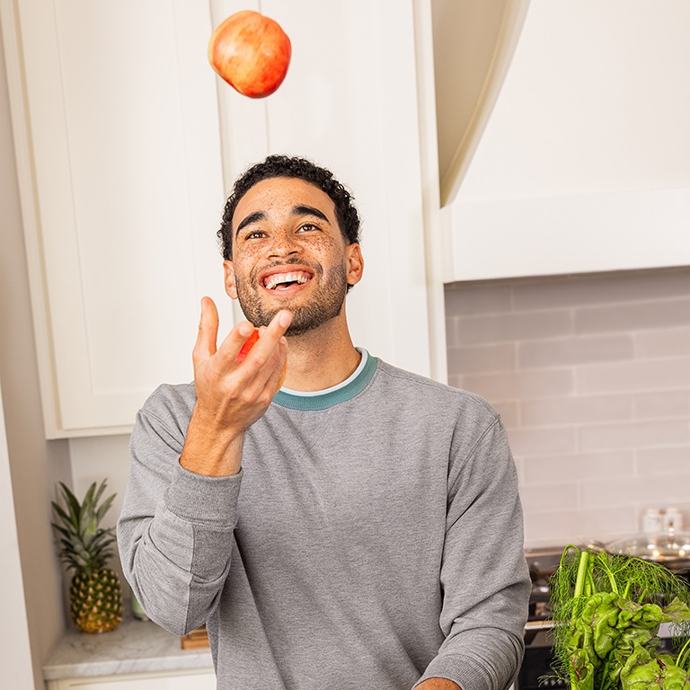 A smiling man in a white kitchen juggles apples. Other produce is visible on the counters.