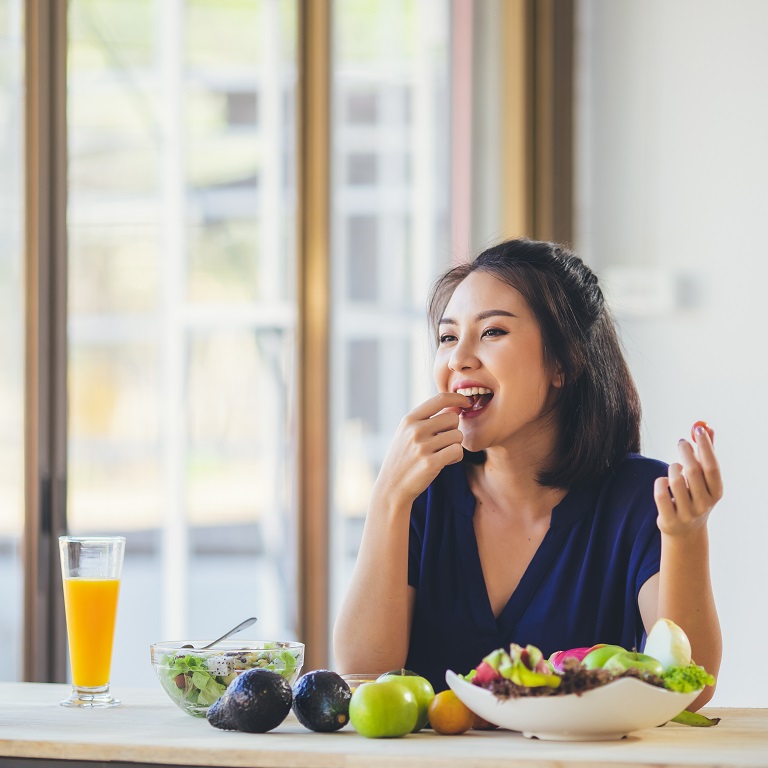 Une femme souriante mange un repas composé de salade et de fruits. D’autres fruits et légumes et un verre de jus d’orange se trouvent à proximité.