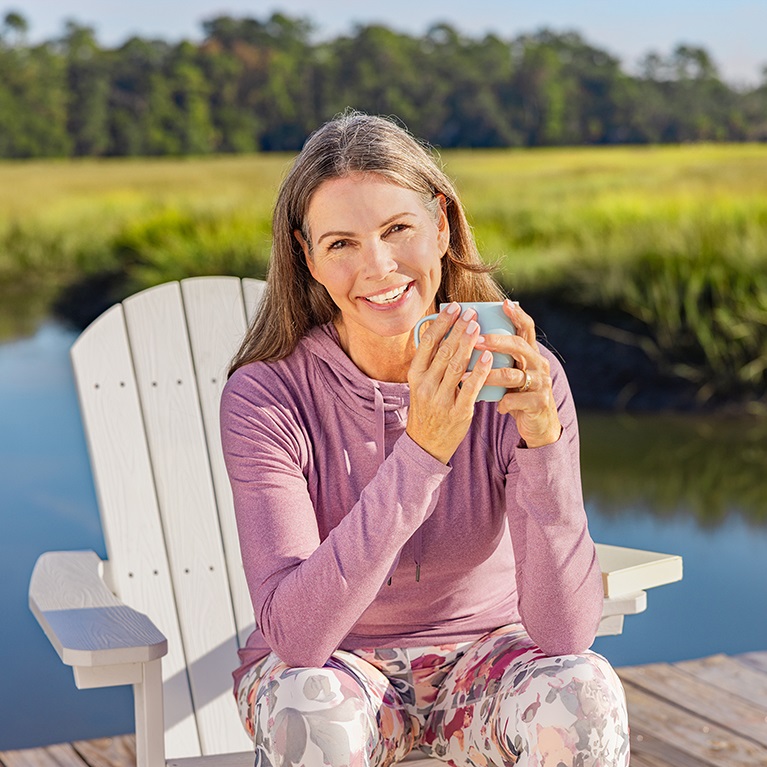 Une femme souriante assise dans un fauteuil Adirondak sur un quai tient une tasse de café ou de thé. Une étendue d’eau, un champ vert et des arbres sont visibles en arrière-plan.