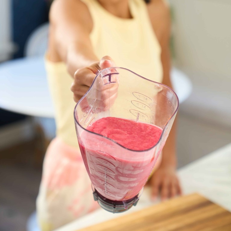A woman wearing a yellow tank top holds a blender containing a tropical berry smoothie over a kitchen counter.