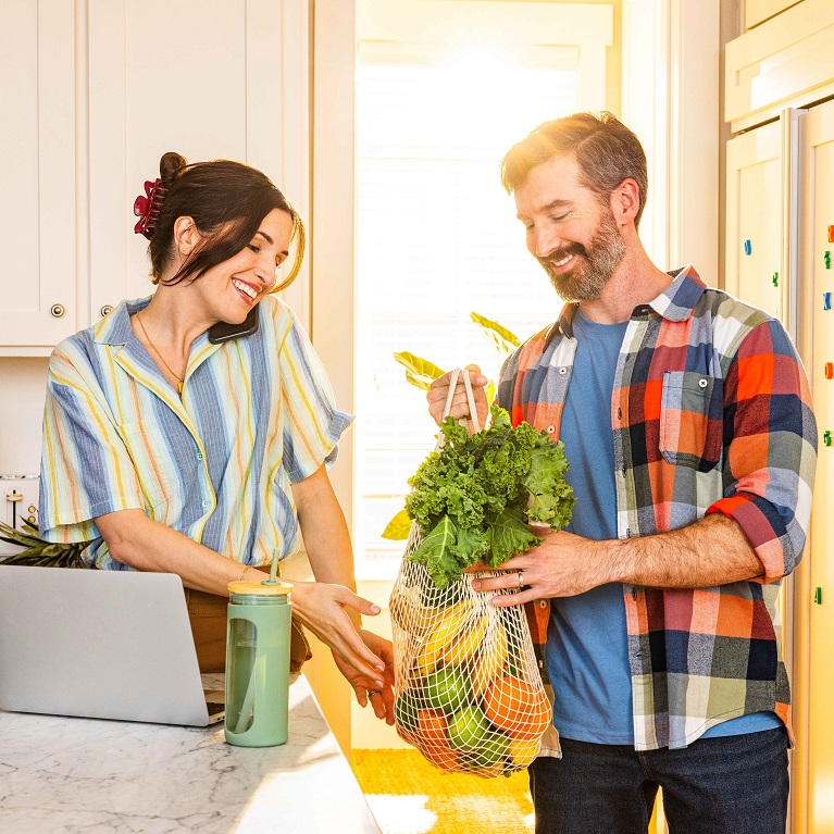 A smiling man hands a woman a mesh back of fresh produce. The woman is smiling while talking on the phone in front of a laptop on a kitchen counter.