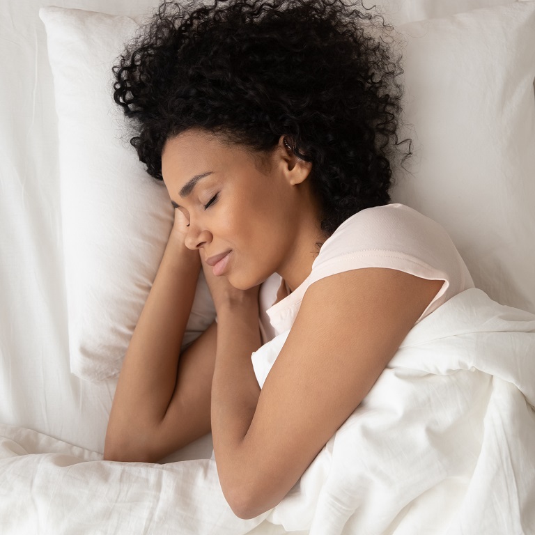 A woman with dark curly hair sleeps on her side in a bed with white bedding. Her head rests on her hands.