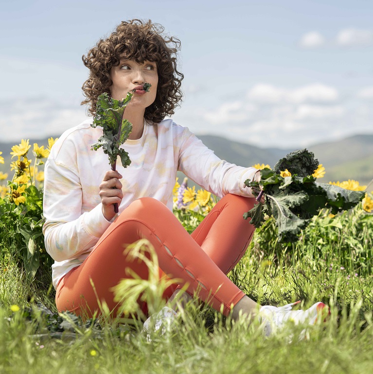 Une femme assise près d’un champ de fleurs jaunes déguste avec plaisir du chou frisé vert.