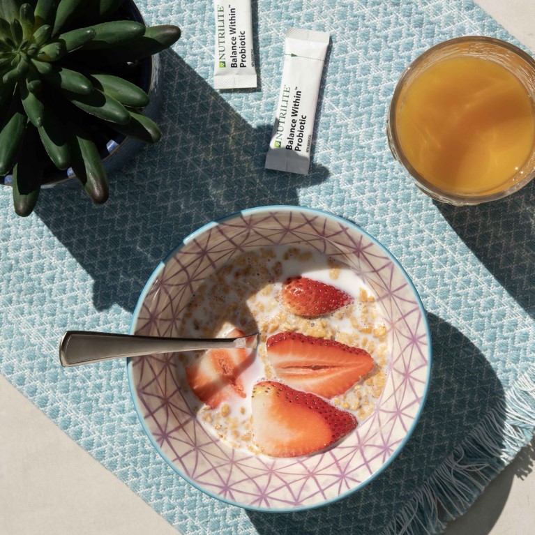 Plusieurs articles sont posés sur un napperon bleu au soleil, dont une plante succulente, des céréales avec du lait et des fraises, du jus d’orange et des probiotiques Balance Within Nutrilite.