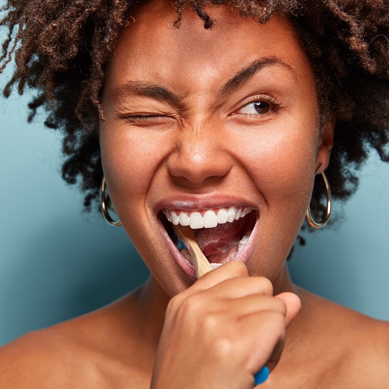 Une femme souriante devant un fond bleu se brosse les dents. Son œil droit est fermé alors que son sourcil gauche est levé de manière excentrique.