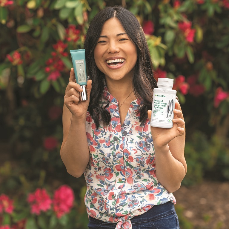 A smiling woman holds a tube of Artistry Skin Nutrition Renewing Reactivation Day Cream and a bottle of Nutrilite Balanced Health Omega.